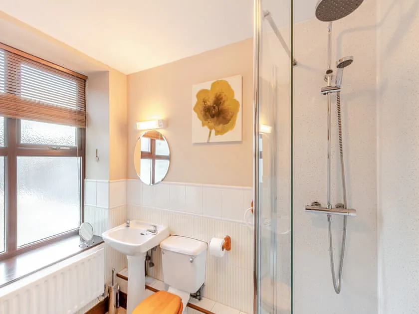 Property for sale by Auction A bright, modern bathroom in The Old Drapery Apartments, 12 Main St, Haltwhistle for sale by auction. It features a glass-enclosed shower with a rainfall showerhead, a white sink with a round mirror above, a toilet with a wooden seat, and framed yellow flower artwork. Frosted windows let in natural light. Presented by Palace Auctions