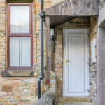 Property for sale by Auction The Old Drapery Apartments at 12 Main St, Haltwhistle, for sale by auction, feature a charming stone exterior with a white door and wooden canopy. Beside the door is a window adorned with lace curtains, and a gutter pipe runs along the wall. This quaint scene appears to be the side entrance of a house. Presented by Palace Auctions