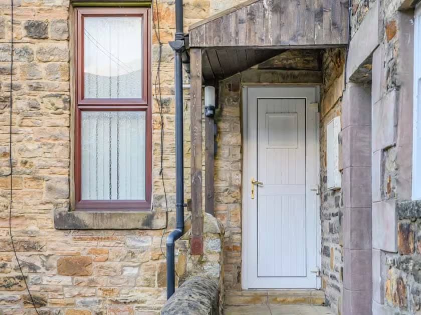 Property for sale by Auction The Old Drapery Apartments at 12 Main St, Haltwhistle, for sale by auction, feature a charming stone exterior with a white door and wooden canopy. Beside the door is a window adorned with lace curtains, and a gutter pipe runs along the wall. This quaint scene appears to be the side entrance of a house. Presented by Palace Auctions