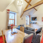Property for sale by Auction A cozy living and dining area at The Old Drapery Apartments, 12 Main St, Haltwhistle. Featuring wooden beams on a high ceiling, it includes a wooden dining table, chairs, a black sofa, wall-mounted TV, and artwork. Red carpet, potted plants, and large windows with curtains complete the inviting atmosphere. For sale by auction. Presented by Palace Auctions