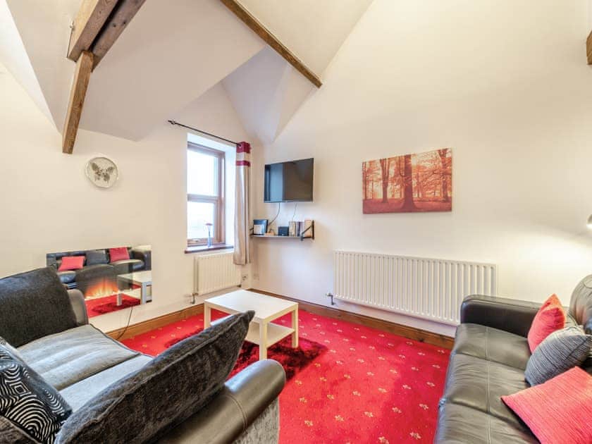 Property for sale by Auction A cozy living room with red carpet, two black leather sofas, and a white coffee table. The room features a wall-mounted flat-screen TV, a fireplace with a mirror above it, and a window with curtains. Ceiling beams are visible in this charming unit at The Old Drapery Apartments, 12 Main St Haltwhistle for sale by auction. Presented by Palace Auctions
