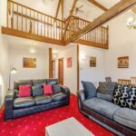Property for sale by Auction A cozy living room at The Old Drapery Apartments, 12 Main St, Haltwhistle, features two sofas, a coffee table, and a vibrant red carpet. The ceiling is high with exposed wooden beams and a mezzanine level overlooks the space. Warm lighting and landscape artwork decorate the white walls, creating a welcoming atmosphere. For sale by auction. Presented by Palace Auctions