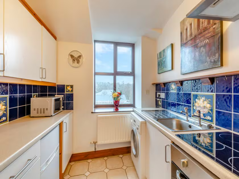 Property for sale by Auction A compact kitchen in The Old Drapery Apartments, 12 Main St, Haltwhistle for sale by auction features white cabinets, a stainless steel sink, a washing machine, and a microwave. Blue tiled walls with floral patterns and a butterfly wall clock add charm. A window above the radiator holds a vase of flowers on the sill. Presented by Palace Auctions