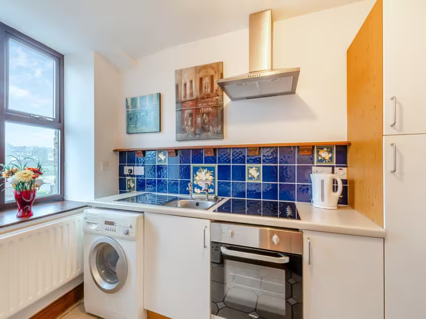 Property for sale by Auction A modern kitchen at The Old Drapery Apartments, 12 Main St, Haltwhistle, for sale by auction. Features a white countertop, electric stove and oven. There’s a blue-tiled backsplash with floral designs, two framed pictures above, and a washing machine by the sink. A white kettle and vase with flowers are on the counter. Large window in the background. Presented by Palace Auctions