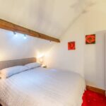 Property for sale by Auction A cozy bedroom in The Old Drapery Apartments, 12 Main St, Haltwhistle, features a double bed with a white comforter and two bedside tables with lamps. Above the bed is a wooden beam. The walls are adorned with two red rose paintings, and the room is illuminated with warm lighting. For sale by auction. Presented by Palace Auctions