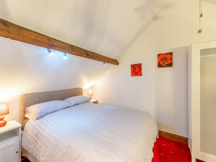 Property for sale by Auction A cozy bedroom in The Old Drapery Apartments, 12 Main St, Haltwhistle, features a double bed with a white comforter and two bedside tables with lamps. Above the bed is a wooden beam. The walls are adorned with two red rose paintings, and the room is illuminated with warm lighting. For sale by auction. Presented by Palace Auctions