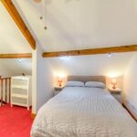 Property for sale by Auction A cozy attic bedroom at The Old Drapery Apartments, 12 Main St Haltwhistle, for sale by auction. The room features a double bed beneath a sloped ceiling with exposed wooden beams, flanked by nightstands with red lamps. A white dresser sits near the railing, and the room has red carpeting and soft, warm lighting. Presented by Palace Auctions