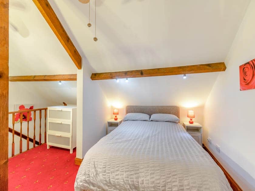 Property for sale by Auction A cozy attic bedroom at The Old Drapery Apartments, 12 Main St Haltwhistle, for sale by auction. The room features a double bed beneath a sloped ceiling with exposed wooden beams, flanked by nightstands with red lamps. A white dresser sits near the railing, and the room has red carpeting and soft, warm lighting. Presented by Palace Auctions