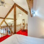 Property for sale by Auction A cozy bedroom loft at The Old Drapery Apartments, 12 Main St, Haltwhistle. Featuring wooden beams, a double bed with a white bedspread, red carpeted flooring, and a wooden rocking chair on the landing. Ceiling fans and a chandelier create a warm and inviting atmosphere. For sale by auction. Presented by Palace Auctions