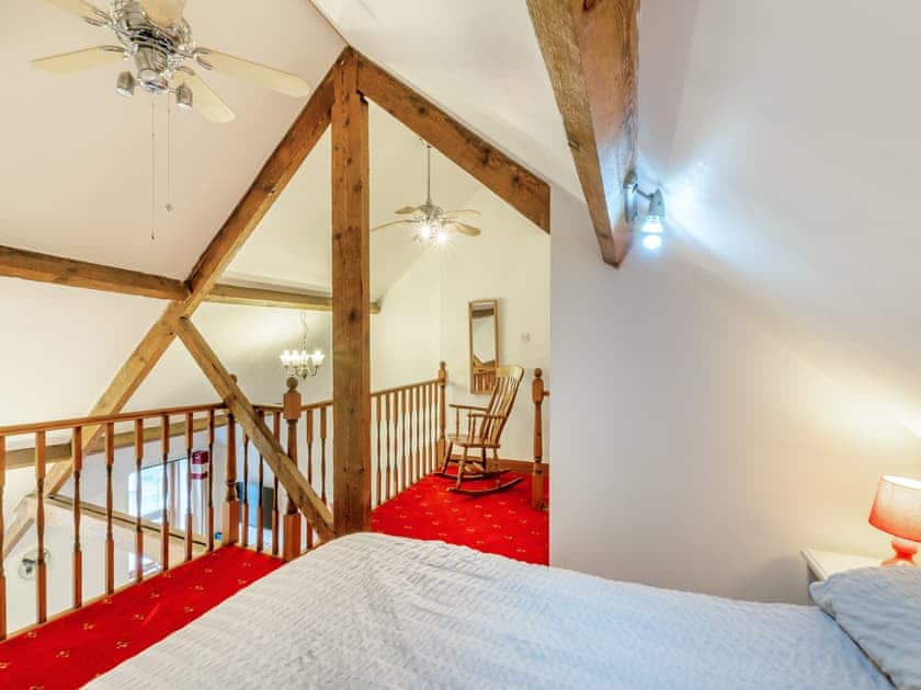 Property for sale by Auction A cozy bedroom loft at The Old Drapery Apartments, 12 Main St, Haltwhistle. Featuring wooden beams, a double bed with a white bedspread, red carpeted flooring, and a wooden rocking chair on the landing. Ceiling fans and a chandelier create a warm and inviting atmosphere. For sale by auction. Presented by Palace Auctions