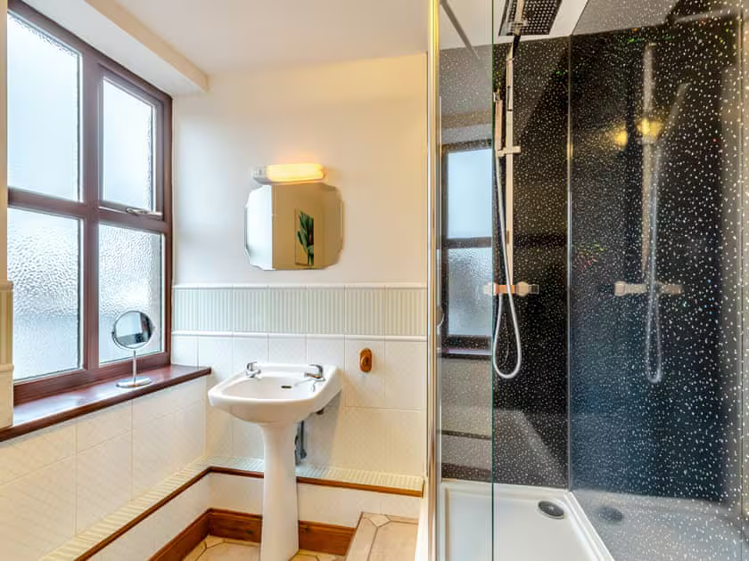 Property for sale by Auction A well-lit bathroom at The Old Drapery Apartments, 12 Main St Haltwhistle for sale by auction features a frosted window, white-tiled walls, and wood accents. Highlights include a sink with a small mirror above it, a round vanity mirror on the windowsill, and a glass-enclosed shower with dark, textured wall tiles. Presented by Palace Auctions
