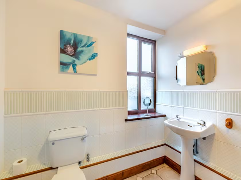 Property for sale by Auction Located in The Old Drapery Apartments, 12 Main St, Haltwhistle, this clean bathroom boasts white walls and tile accents. It features a white toilet, a pedestal sink with a wall-mounted mirror above it, a small window, and a painting of a flower on the wall. For sale by auction. Presented by Palace Auctions
