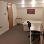 Property for sale by Auction A small, simply furnished bedroom with a bed, nightstand, desk, chair, and a colorful abstract painting on the wall. The room has beige walls, a wooden door, and a carpeted floor. Two ceiling lights illuminate the space. Presented by Palace Auctions