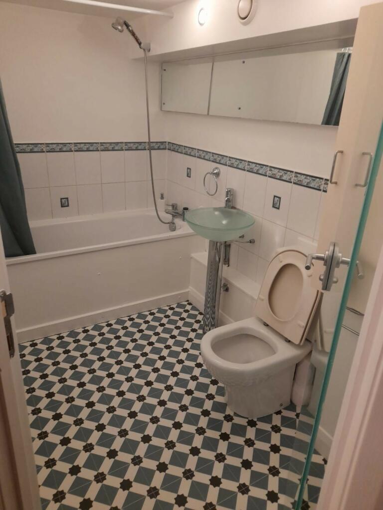 Property for sale by Auction A bathroom featuring a bathtub with a showerhead, a green pedestal sink, a toilet with the lid open, and a floor with black and white checkered tiles. The walls are white with decorative blue and white tiles lining the middle. A large mirror is above the sink and tub. Presented by Palace Auctions