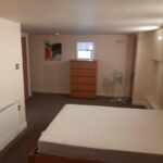 Property for sale by Auction A minimalist bedroom with a mattress on a wooden bed frame, a tall wooden dresser, a small window, a fan on a stand, and abstract paintings on the walls. The room has beige walls and a carpeted floor. Presented by Palace Auctions