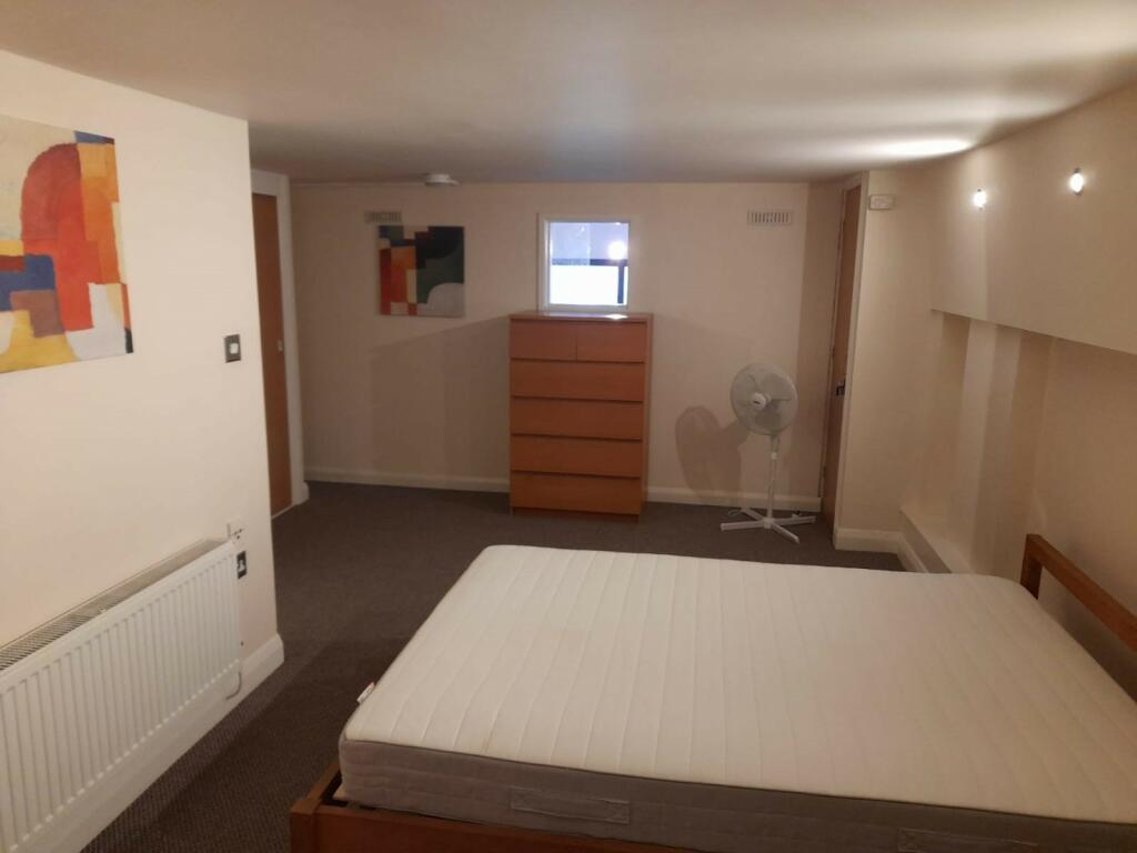Property for sale by Auction A minimalist bedroom with a mattress on a wooden bed frame, a tall wooden dresser, a small window, a fan on a stand, and abstract paintings on the walls. The room has beige walls and a carpeted floor. Presented by Palace Auctions