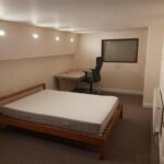 Property for sale by Auction A minimalist room with a single bed featuring a white mattress on a wooden frame. Adjacent to the bed is a wooden desk with a black office chair. The room has recessed ceiling lights, beige walls, and a carpeted floor. A small window and a radiator are visible. Presented by Palace Auctions