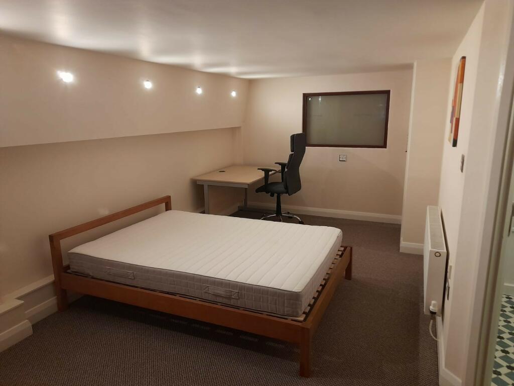 Property for sale by Auction A minimalist room with a single bed featuring a white mattress on a wooden frame. Adjacent to the bed is a wooden desk with a black office chair. The room has recessed ceiling lights, beige walls, and a carpeted floor. A small window and a radiator are visible. Presented by Palace Auctions