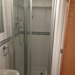 Property for sale by Auction A small bathroom featuring a corner shower with glass doors. The shower has white tiled walls with a decorative tile stripe running horizontally. To the left of the shower is a white sink with a metal towel ring attached to the wall. A wooden door is partially visible on the right. Presented by Palace Auctions