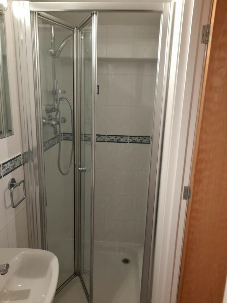 Property for sale by Auction A small bathroom featuring a corner shower with glass doors. The shower has white tiled walls with a decorative tile stripe running horizontally. To the left of the shower is a white sink with a metal towel ring attached to the wall. A wooden door is partially visible on the right. Presented by Palace Auctions