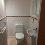 Property for sale by Auction A compact bathroom with a white toilet placed near the back wall, a small white pedestal sink on the right side, and a radiator on the left. The walls are tiled halfway up with a decorative border, and a mirrored cabinet is above the sink. Presented by Palace Auctions