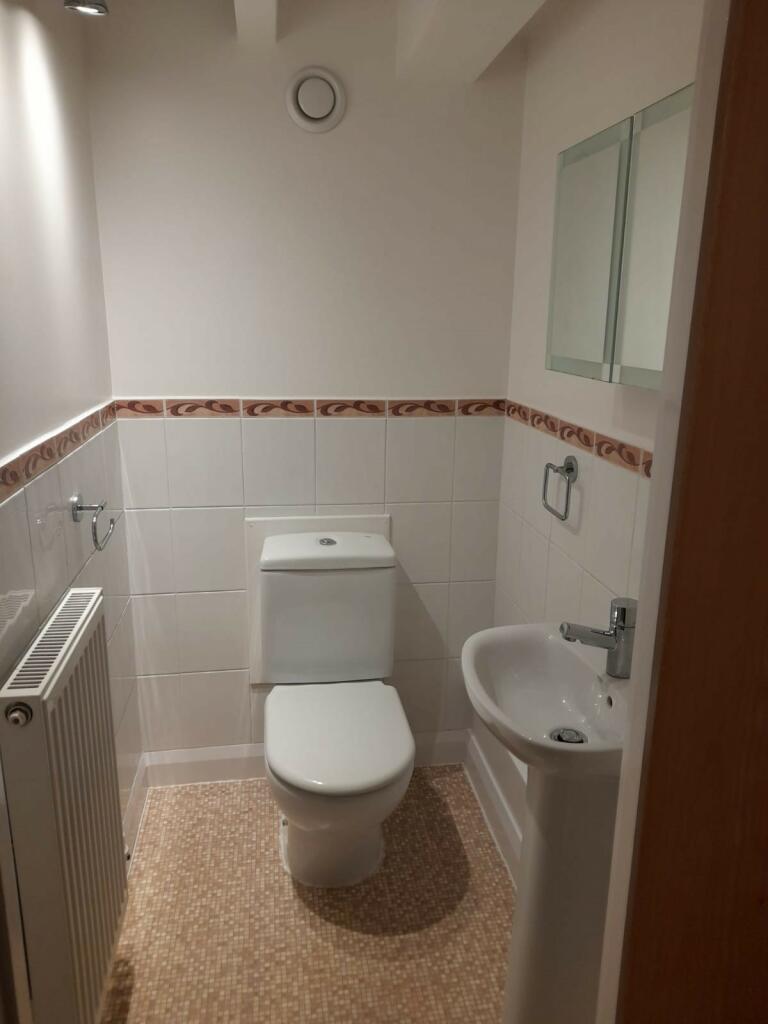 Property for sale by Auction A compact bathroom with a white toilet placed near the back wall, a small white pedestal sink on the right side, and a radiator on the left. The walls are tiled halfway up with a decorative border, and a mirrored cabinet is above the sink. Presented by Palace Auctions