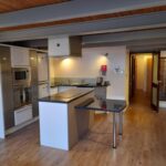 Property for sale by Auction A modern kitchen with wooden flooring and an exposed wooden ceiling. It features white cabinets, stainless steel appliances including a microwave and oven, a central island with a black countertop, and an attached glass table. A door is open in the background. Presented by Palace Auctions