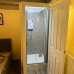 Property for sale by Auction A small bathroom with wooden ceiling features a shower cabin with a clear glass door and a white electric shower unit. The room also has a beige wall, radiator, and part of a gray upholstered bed can be seen. The flooring is wood pattern laminate. Presented by Palace Auctions