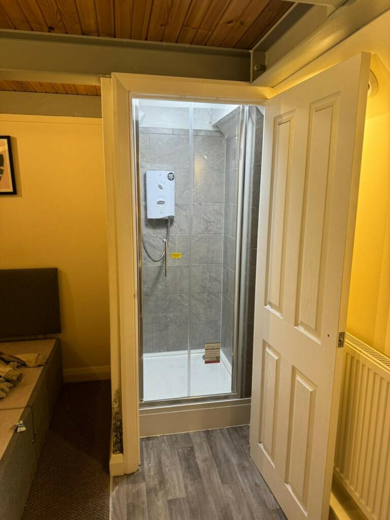 Property for sale by Auction A small bathroom with wooden ceiling features a shower cabin with a clear glass door and a white electric shower unit. The room also has a beige wall, radiator, and part of a gray upholstered bed can be seen. The flooring is wood pattern laminate. Presented by Palace Auctions