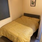 Property for sale by Auction A small bedroom with a bed featuring a yellow and white striped duvet. The bed has a gray headboard and is positioned against the walls in the corner of the room. A framed picture with a leaf design hangs on the wall above the bed. Carpeting covers part of the floor. Presented by Palace Auctions