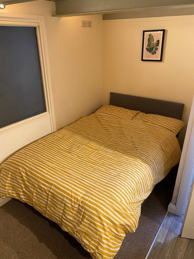 Property for sale by Auction A small bedroom with a bed featuring a yellow and white striped duvet. The bed has a gray headboard and is positioned against the walls in the corner of the room. A framed picture with a leaf design hangs on the wall above the bed. Carpeting covers part of the floor. Presented by Palace Auctions
