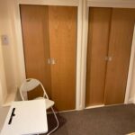 Property for sale by Auction A room with two wooden sliding closet doors. In front of the closets, there is a white folding chair and a small white desk with a black handle. The floor is carpeted, and part of a bed with a yellow and white striped blanket is visible in the corner. Presented by Palace Auctions