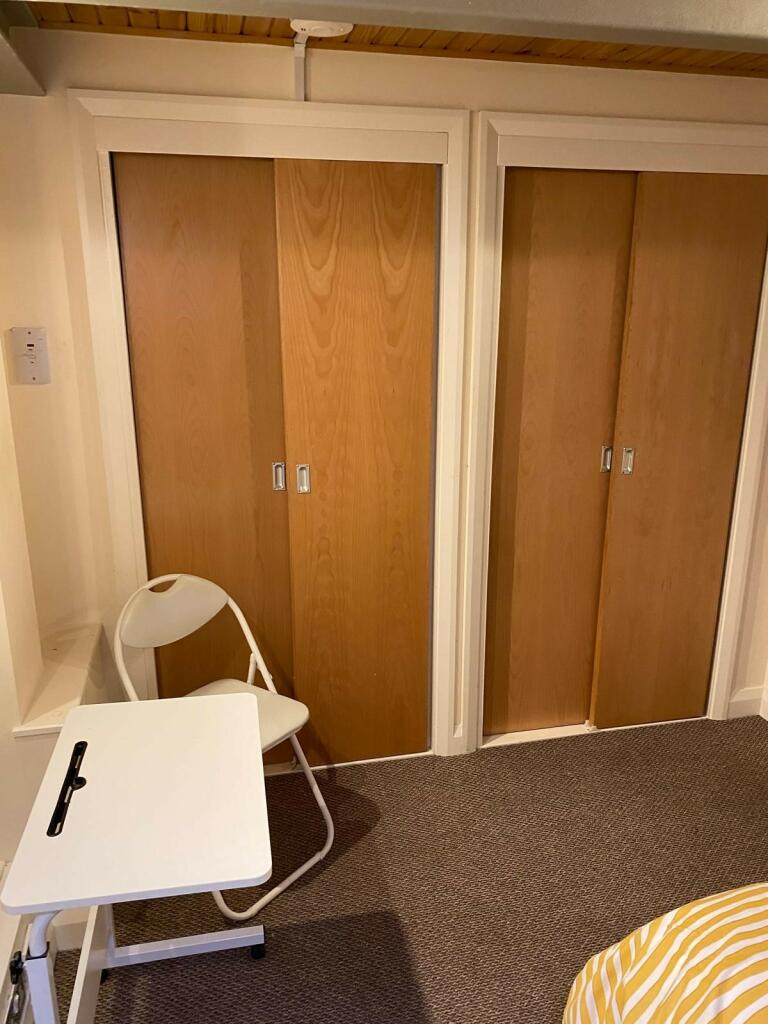 Property for sale by Auction A room with two wooden sliding closet doors. In front of the closets, there is a white folding chair and a small white desk with a black handle. The floor is carpeted, and part of a bed with a yellow and white striped blanket is visible in the corner. Presented by Palace Auctions