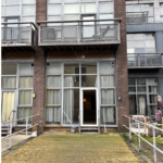 Property for sale by Auction A building facade featuring large glass windows and balconies with wooden railings. The ground level has a wide doorway with an extended wooden ramp for accessibility. The building exterior shows signs of wear, with some plants and outdoor furniture on the side. Presented by Palace Auctions
