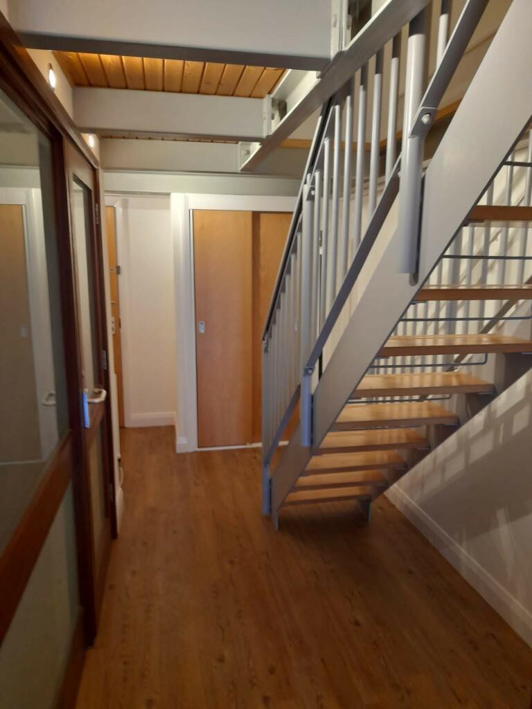 Property for sale by Auction A hallway with wooden flooring and white walls features a modern staircase with metallic railings and wooden steps on the right. On the left side, there are glass doors with wooden frames. A wooden ceiling adds warmth to the space. Presented by Palace Auctions