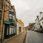 Property for sale by Auction A narrow street lined with historic buildings, including The Old Drapery Apartments at 12 Main St Haltwhistle for sale by auction. The exteriors, a mix of stone and brick in yellows, beiges, and whites, feature unique windows. Parked cars and signs wet from rain are visible under the overcast sky. Presented by Palace Auctions