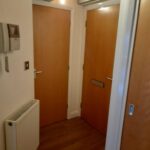 Property for sale by Auction A hallway with two closed wooden doors facing each other, a wall-mounted intercom system, and a radiator. The hardwood floor and light-colored walls give the space a warm feel. A portion of a third doorway is visible on the right side of the image. Presented by Palace Auctions