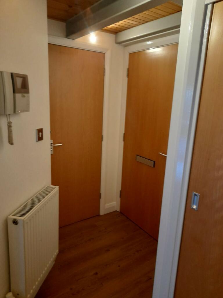 Property for sale by Auction A hallway with two closed wooden doors facing each other, a wall-mounted intercom system, and a radiator. The hardwood floor and light-colored walls give the space a warm feel. A portion of a third doorway is visible on the right side of the image. Presented by Palace Auctions