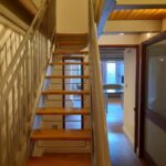 Property for sale by Auction A narrow wooden staircase with metal railings ascends to an upper floor. The area features wooden flooring and ceiling. In the background, there is a sunlit room with a large window and a table, accessible through a doorway. Presented by Palace Auctions
