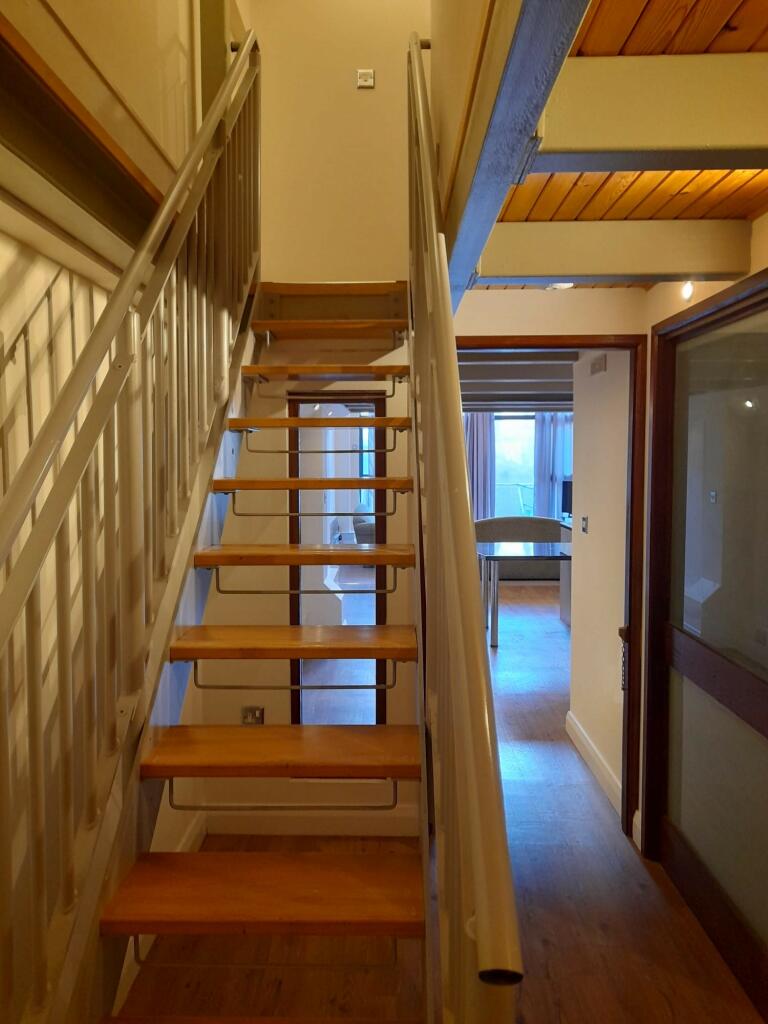 Property for sale by Auction A narrow wooden staircase with metal railings ascends to an upper floor. The area features wooden flooring and ceiling. In the background, there is a sunlit room with a large window and a table, accessible through a doorway. Presented by Palace Auctions