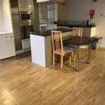 Property for sale by Auction A modern kitchen with wooden flooring, featuring a small fridge, built-in oven and microwave, a transparent glass table with wooden chairs, and a countertop separating the kitchen from the rest of the room. There's also a garbage bin and a fire extinguisher on the wall. Presented by Palace Auctions