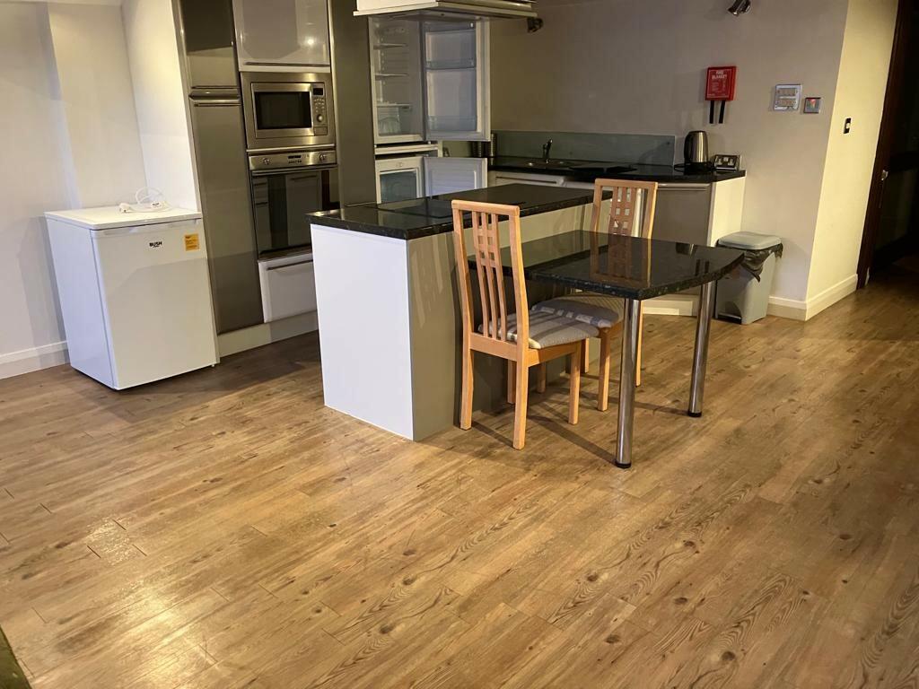 Property for sale by Auction A modern kitchen with wooden flooring, featuring a small fridge, built-in oven and microwave, a transparent glass table with wooden chairs, and a countertop separating the kitchen from the rest of the room. There's also a garbage bin and a fire extinguisher on the wall. Presented by Palace Auctions