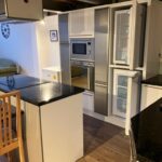 Property for sale by Auction A modern kitchen with wooden flooring features a built-in stainless steel oven, microwave, and refrigerator with the doors open. A black granite-topped island with a wooden chair is in the foreground, and a small bed is visible in the background. Presented by Palace Auctions