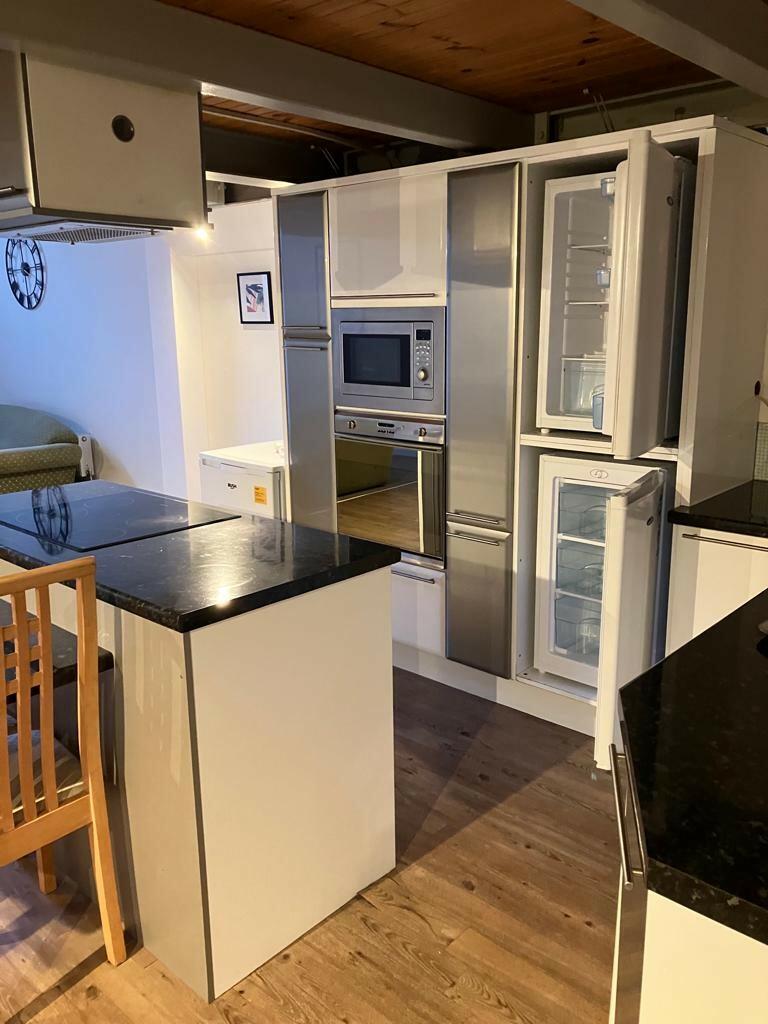 Property for sale by Auction A modern kitchen with wooden flooring features a built-in stainless steel oven, microwave, and refrigerator with the doors open. A black granite-topped island with a wooden chair is in the foreground, and a small bed is visible in the background. Presented by Palace Auctions