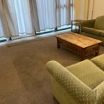Property for sale by Auction A living room with a green couch and matching armchair, both with polka dot upholstery, placed on a carpeted floor. There's a wooden coffee table in front of the couch. The room has large windows with closed white curtains and a glass side table beside the couch. Presented by Palace Auctions