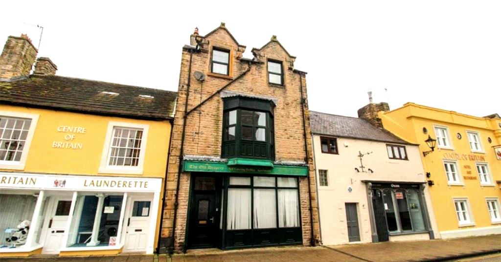 Property for sale by Auction A quaint town street featuring a row of charming buildings. The central building, known as The Old Drapery Apartments, has a brick exterior with a distinctive green sign and an upper-floor bay window. To the left is "Centre of Britain" laundrette, while the adjacent buildings have colorful facades. Presented by Palace Auctions