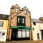 Property for sale by Auction A quaint town street featuring a row of charming buildings. The central building, known as The Old Drapery Apartments, has a brick exterior with a distinctive green sign and an upper-floor bay window. To the left is "Centre of Britain" laundrette, while the adjacent buildings have colorful facades. Presented by Palace Auctions