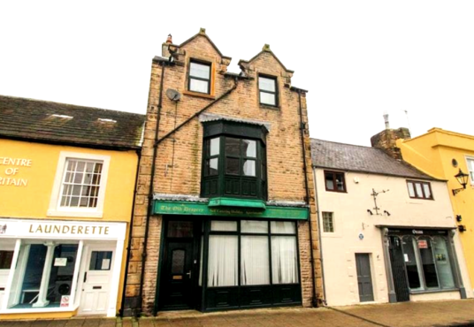 Property for sale by Auction A quaint town street featuring a row of charming buildings. The central building, known as The Old Drapery Apartments, has a brick exterior with a distinctive green sign and an upper-floor bay window. To the left is "Centre of Britain" laundrette, while the adjacent buildings have colorful facades. Presented by Palace Auctions