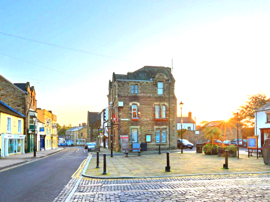 Property for sale by Auction A quaint town square with a historic brick building in the center. The sun sets, casting a warm glow over the scene. Cobblestone streets and a variety of small shops line the area, giving a charming and peaceful atmosphere. Nearby, The Old Drapery Apartments at 12 Main St Haltwhistle are for sale by auction. Presented by Palace Auctions