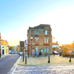 Property for sale by Auction A quaint town square with a historic brick building in the center. The sun sets, casting a warm glow over the scene. Cobblestone streets and a variety of small shops line the area, giving a charming and peaceful atmosphere. Nearby, The Old Drapery Apartments at 12 Main St Haltwhistle are for sale by auction. Presented by Palace Auctions